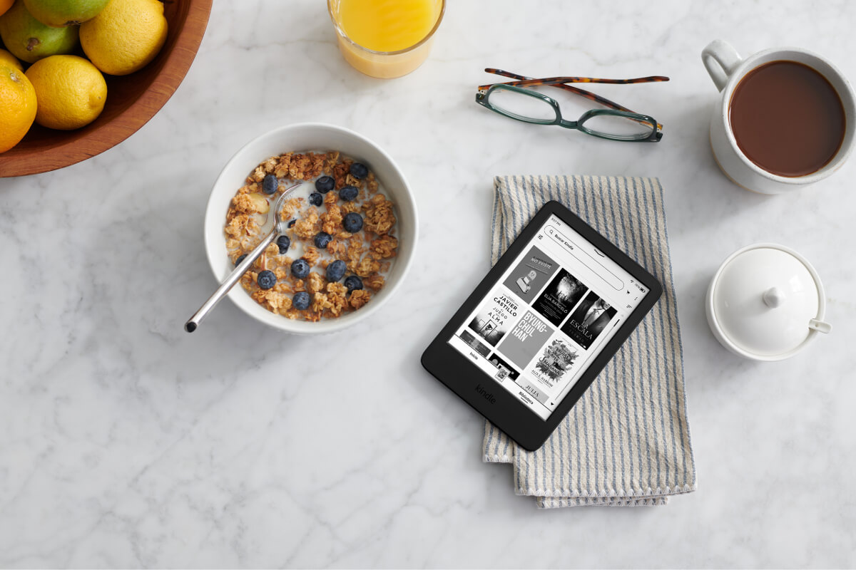 La nueva generación del lector Kindle en negro, acompañada de un desayuno en una mesa.